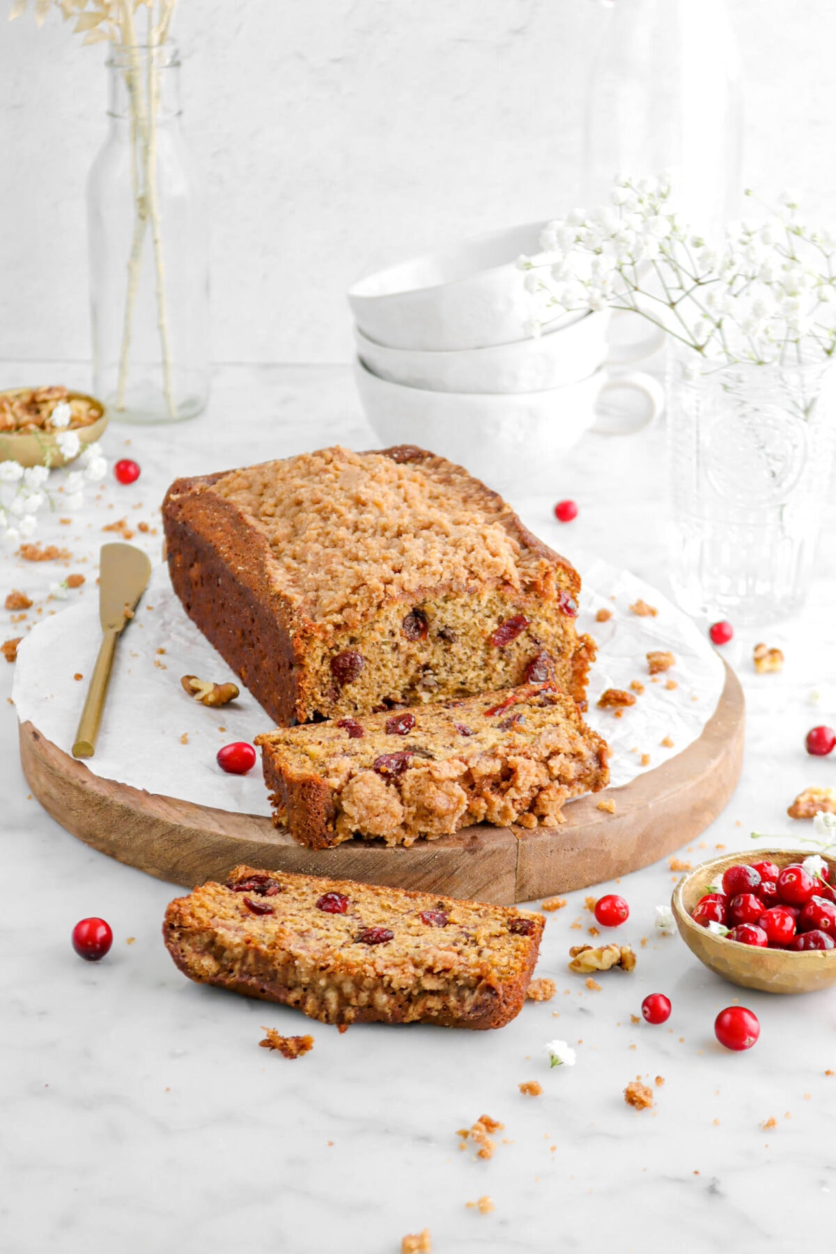 two slices of cranberry walnut banana bread laying in front of loaf on wood board with gold knife beside, fresh cranberries, white flowers, and walnut pieces around.