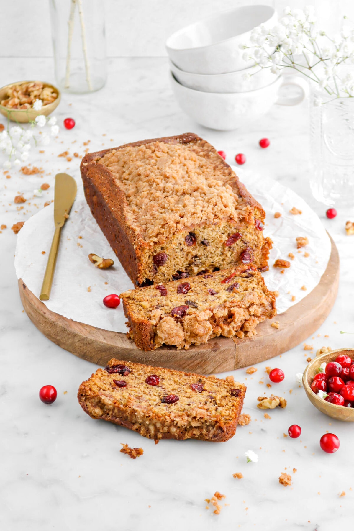 angled shot of banana bread on wood board with two slices laying in front, a gold knife beside, bowl of cranberries in front, and a bowl of walnuts behind.