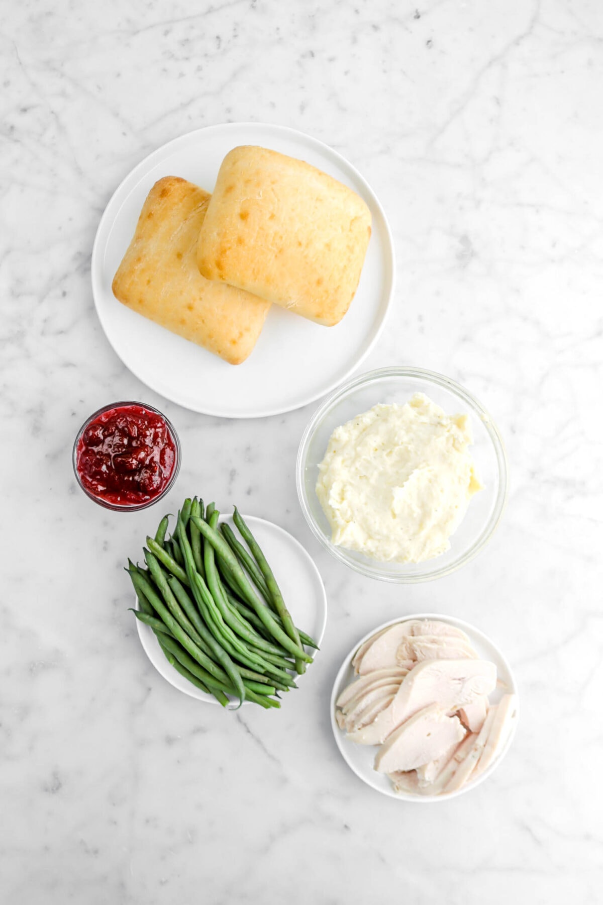 two ciabatta rolls, cranberry sauce, mashed potatoes, green beans, and sliced turkey on marble surface.