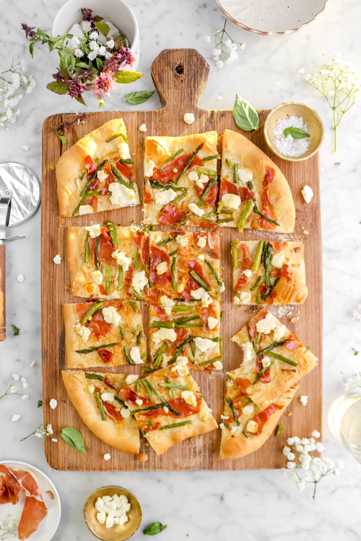 sliced asparagus and prosciutto flatbread on wood board with one slice missing a bite, a gold bowl beside, fresh flowers, and basil leaves around on marble surface.