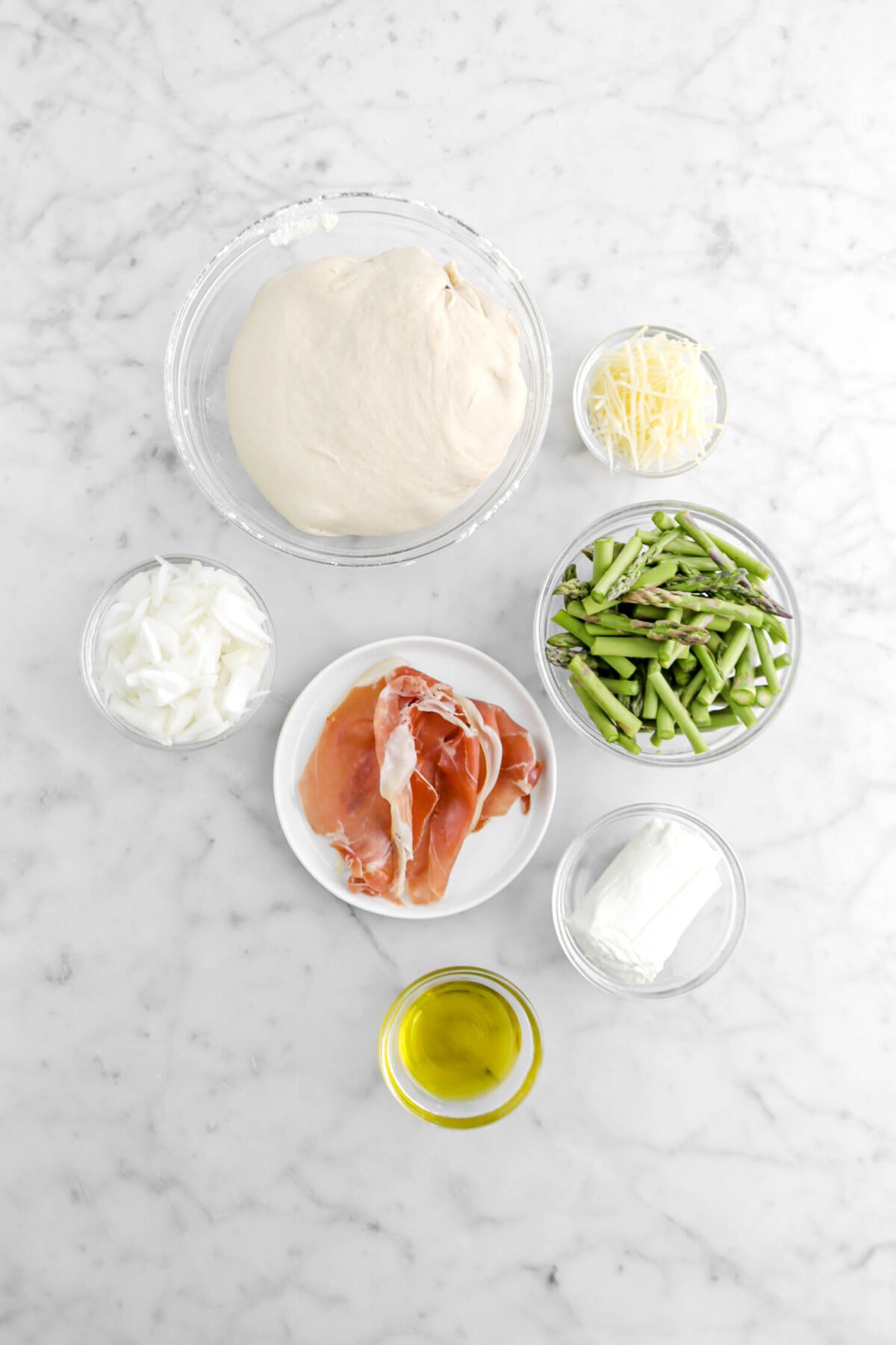 pizza dough, grated parmesan cheese, slices onion, prosciutto, asparagus tops, log of goat cheese, and olive oil on marble surface.