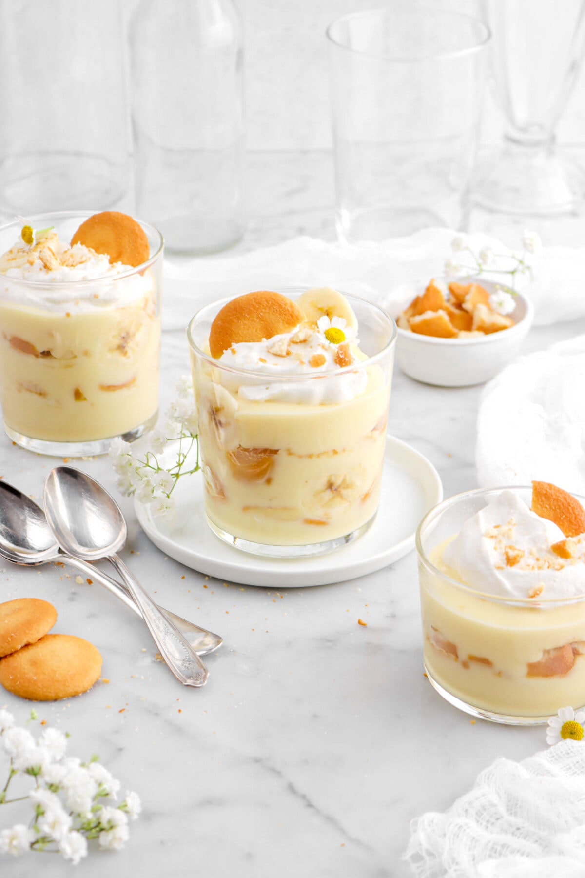 angled shot of three glasses of banana pudding with two spoons beside, white flowers, vanilla wafers, and white cheese cloth around.