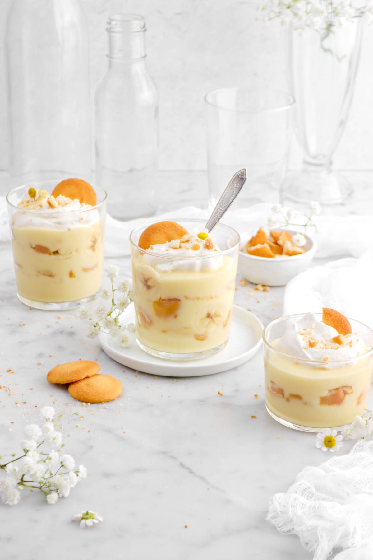 three glasses of banana pudding with spoon dug into middle glass with vanilla wafers and flowers around.
