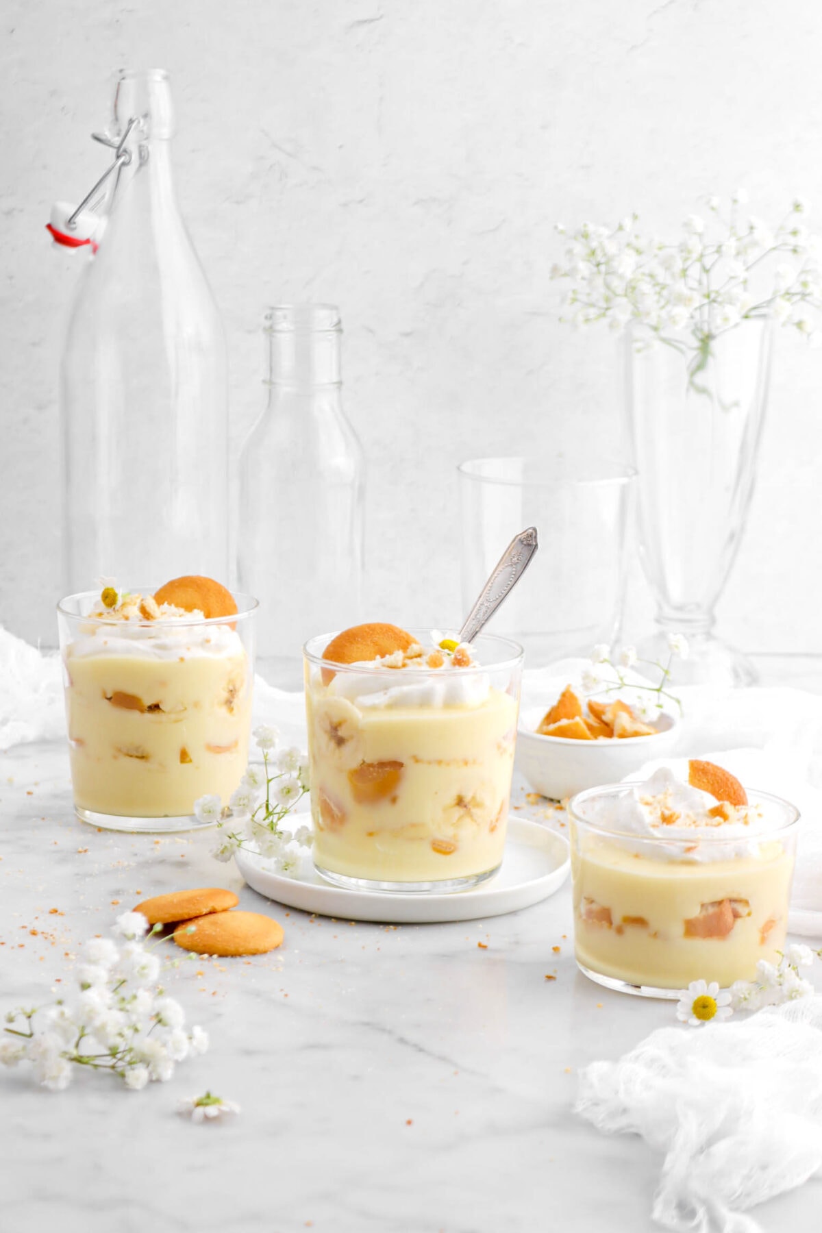 front shot of three glasses of banana pudding with spoon dug inside middle glass with flowers, white cheese cloth, and vanilla wafers around.
