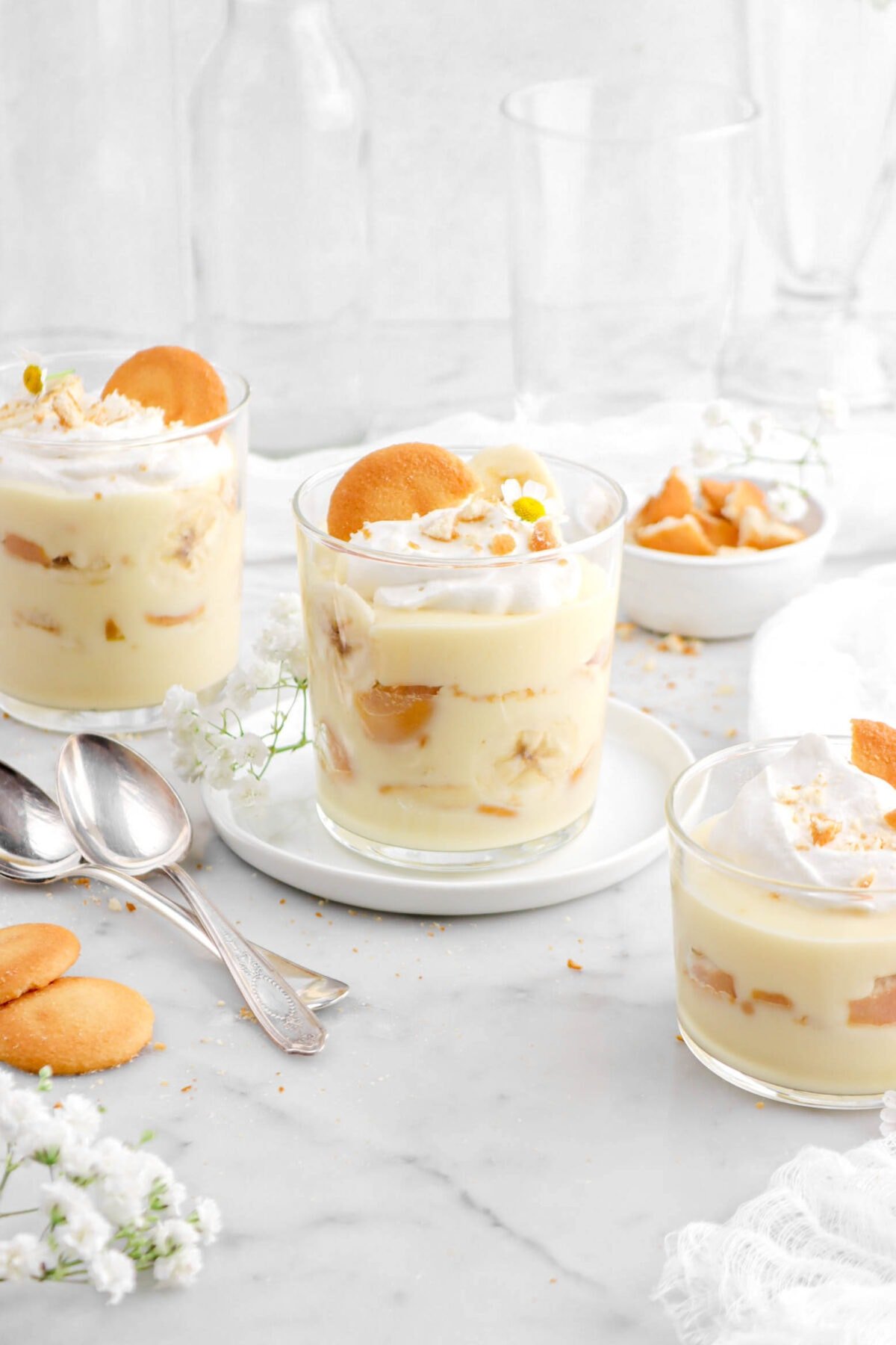 glasses of banana pudding with two spoons beside, vanilla wafers and flowers around on marble surface.