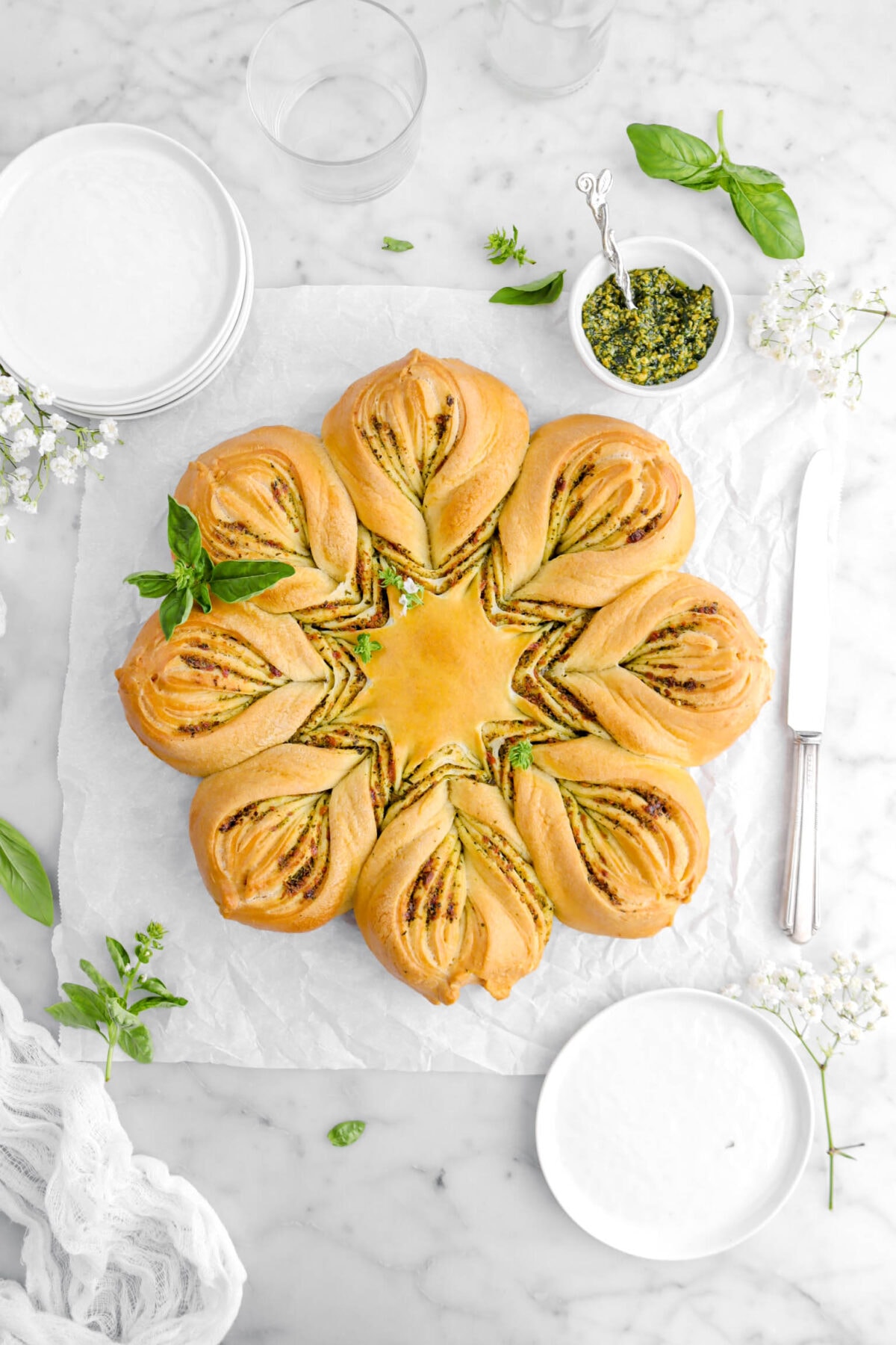 overhead shot of basil pesto star bread with basil, white flowers, and plates around.