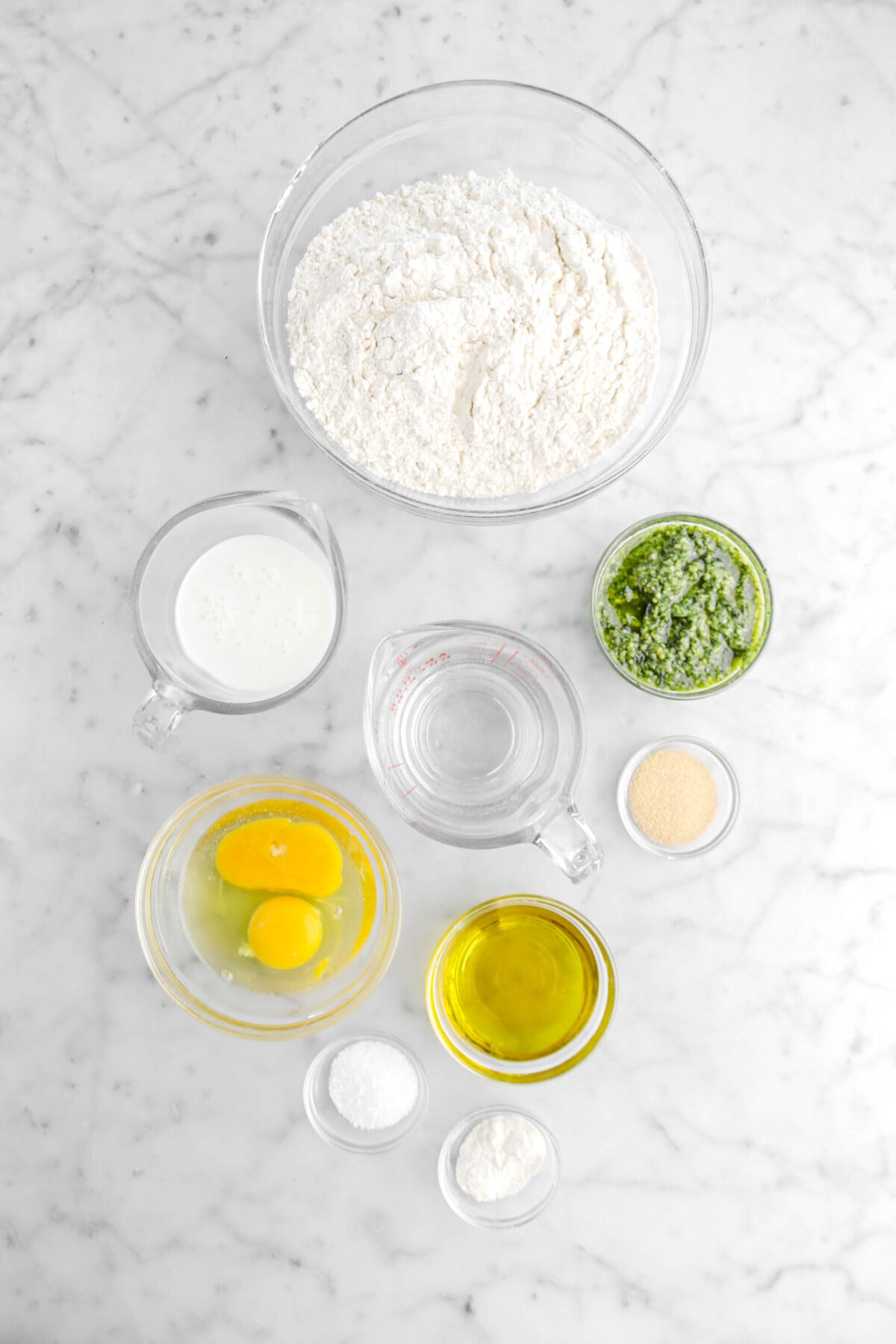 flour, milk, water, basil pesto, eggs, yeast, olive oil salt, and malt powder on marble surface.