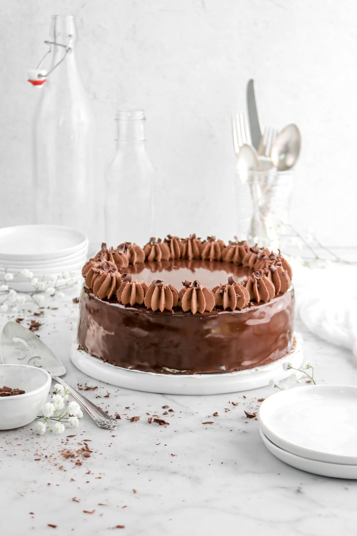 front shot of chocolate mousse cake with mirror glaze on upside down white plate with white flowers and chocolate curls around, stack of white plates, empty glasses, and utensils behind.