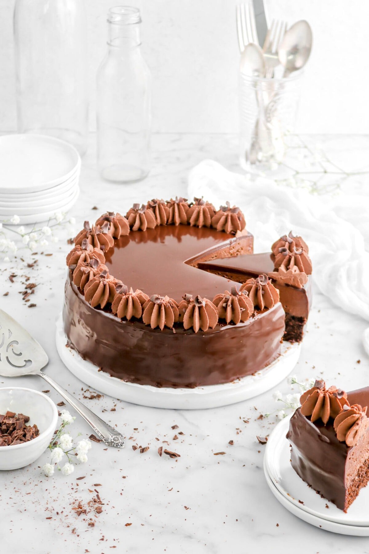 angled shot of sliced chocolate mousse cake with another slice beside on stack of two plates, a cake knife and bowl of chocolate curls beside, white flowers and cheese cloth behind.