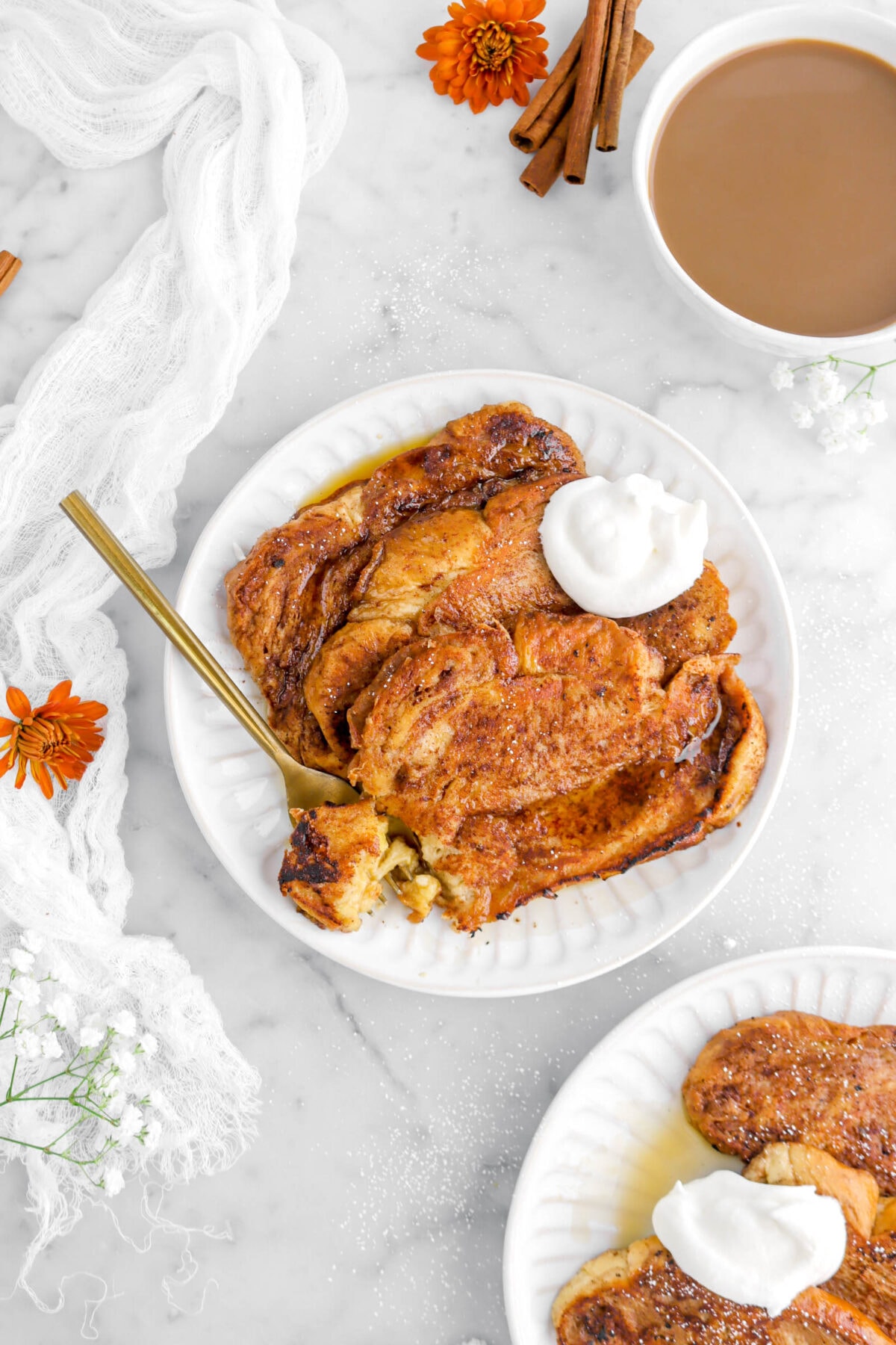 three slices of cinnamon french toast with gold fork beside, a mug of coffee, cinnamon sticks, flowers, a second plate, and white cheese cloth around on marble surface.