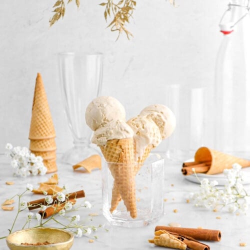 pulled back shot of two cones of cinnamon ice cream in white glass with gold garland above, white flowers, and cinnamon sticks around on marble surface.