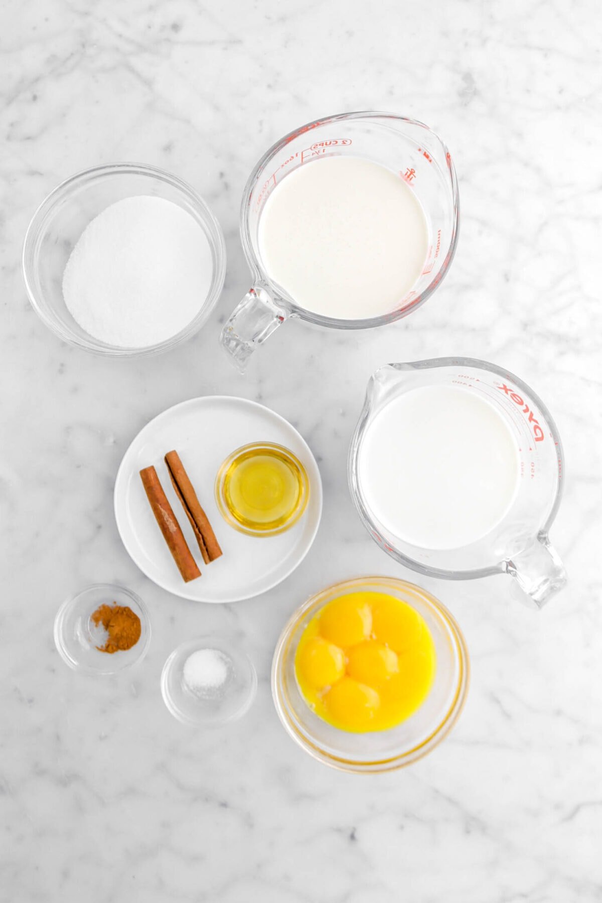 sugar, heavy cream, milk, agave, two cinnamon sticks, ground cinnamon, salt, and egg yolks on marble surface.