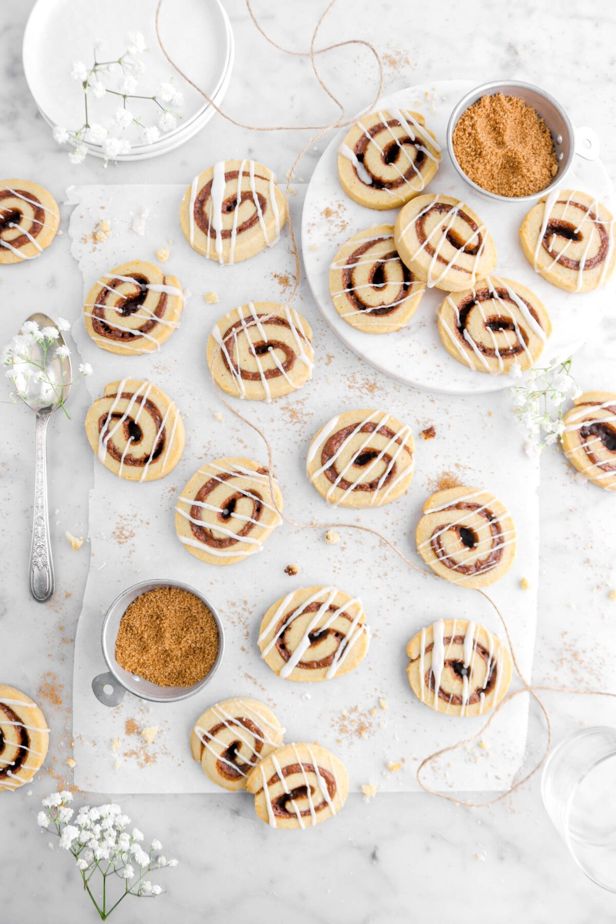 cinnamon roll cookies on parchment paper with marble riser beside with more cookies, two measuring cups of cinnamon sugar, and white flowers around on marble surface.