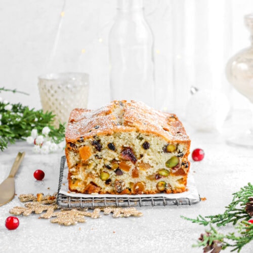 fruit cake on wire tray with gold snowflake ornament underneath, gold knife beside, cranberries, and greenery around with powdered sugar dusted on top of cake.
