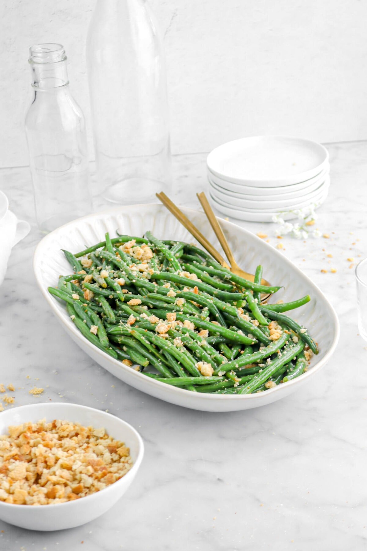 green beans in deep platter with bowl of bread crumbs beside, two empty glasses, and a stack of white plates behind.