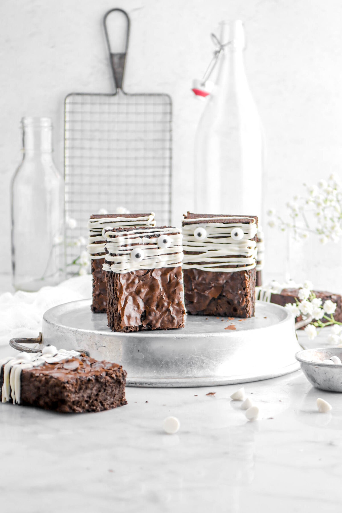 three brownies standing up right on pie plate with wire cooling rack, empty glasses, and flowers behind.