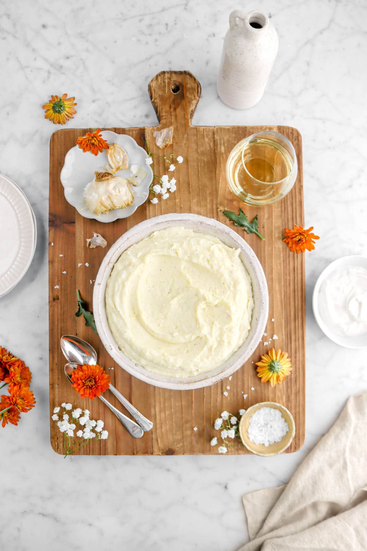 overhead shot of bowl of mashed potatoes on large wood board on a marble surface with flowers, plates, and a bowl of sour cream beside, with an empty vase, and a napkin around wood board.