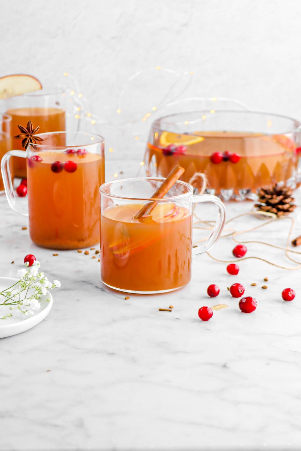 pulled back shot of apple cider in glass mugs with apple slices and cinnamon stick in front mug, punch bowl, small pinecone, fairy lights, and brown twine behind.