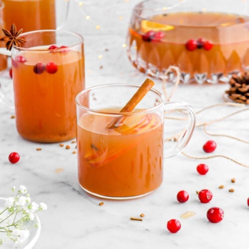 angled photo of three mugs of apple cider on marble surface with punch bowl and fairy lights behind, fresh cranberries and cinnamon chunks around.
