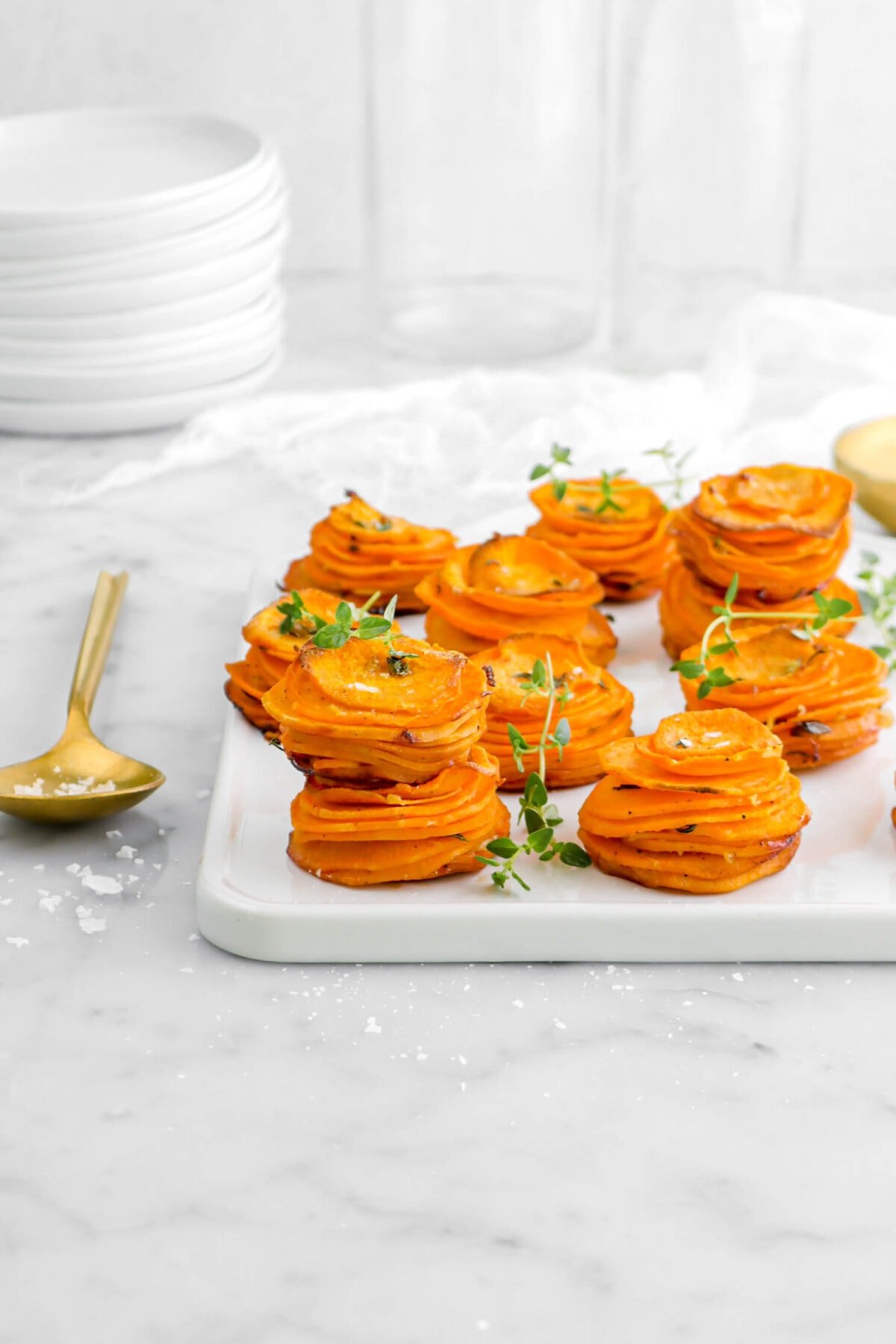 pulled back shot of sweet potato stacks on square white tray with gold spoon beside, flaked salt, and thyme sprigs on marble surface with stack of plates behind.
