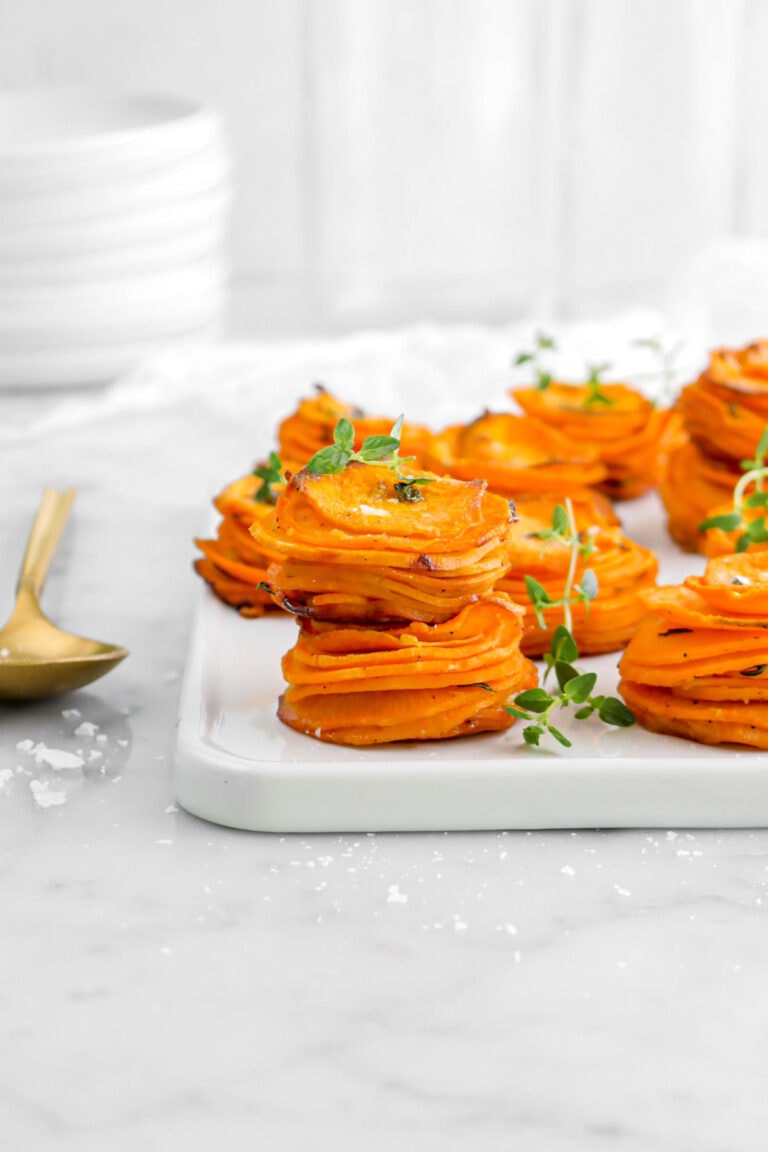 front shot of sweet potato stacks on white square tray with thyme sprigs and flaked salt on top, fold spoon beside, and stack of white plates behind.