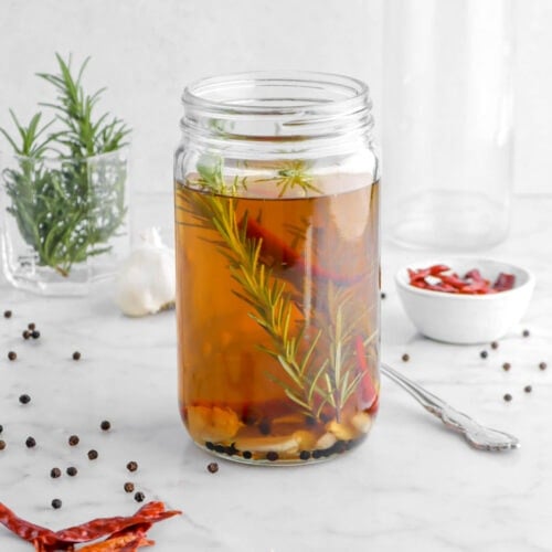angled shot of turkey brine in glass jar with rosemary, garlic, peppercorns, and chilis in brine with the same ingredients on marble surface around jar.