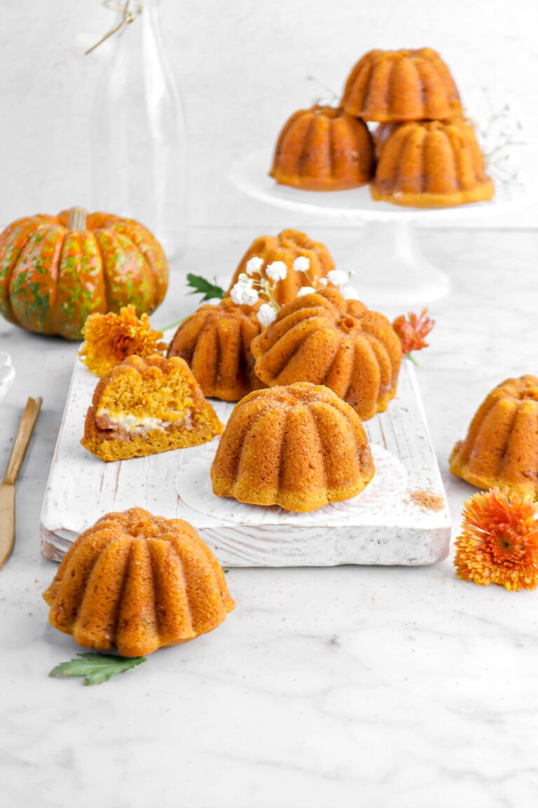 four pumpkin shaped cakes on white board with one cut in half, flowers and three more cakes around, a mini pumpkin, and a cake stand with more cakes behind.