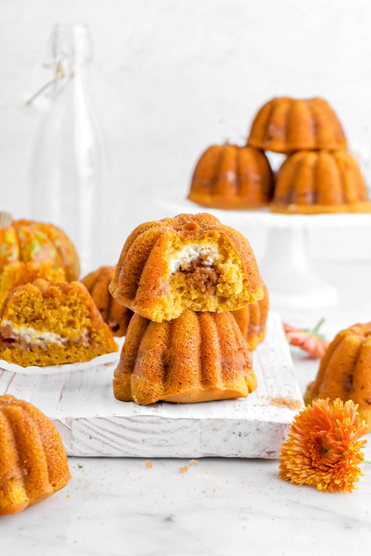 two stacked pumpkin cakes with one missing a bite, more pumpkin cakes behind, and an orange chrysanthemum.
