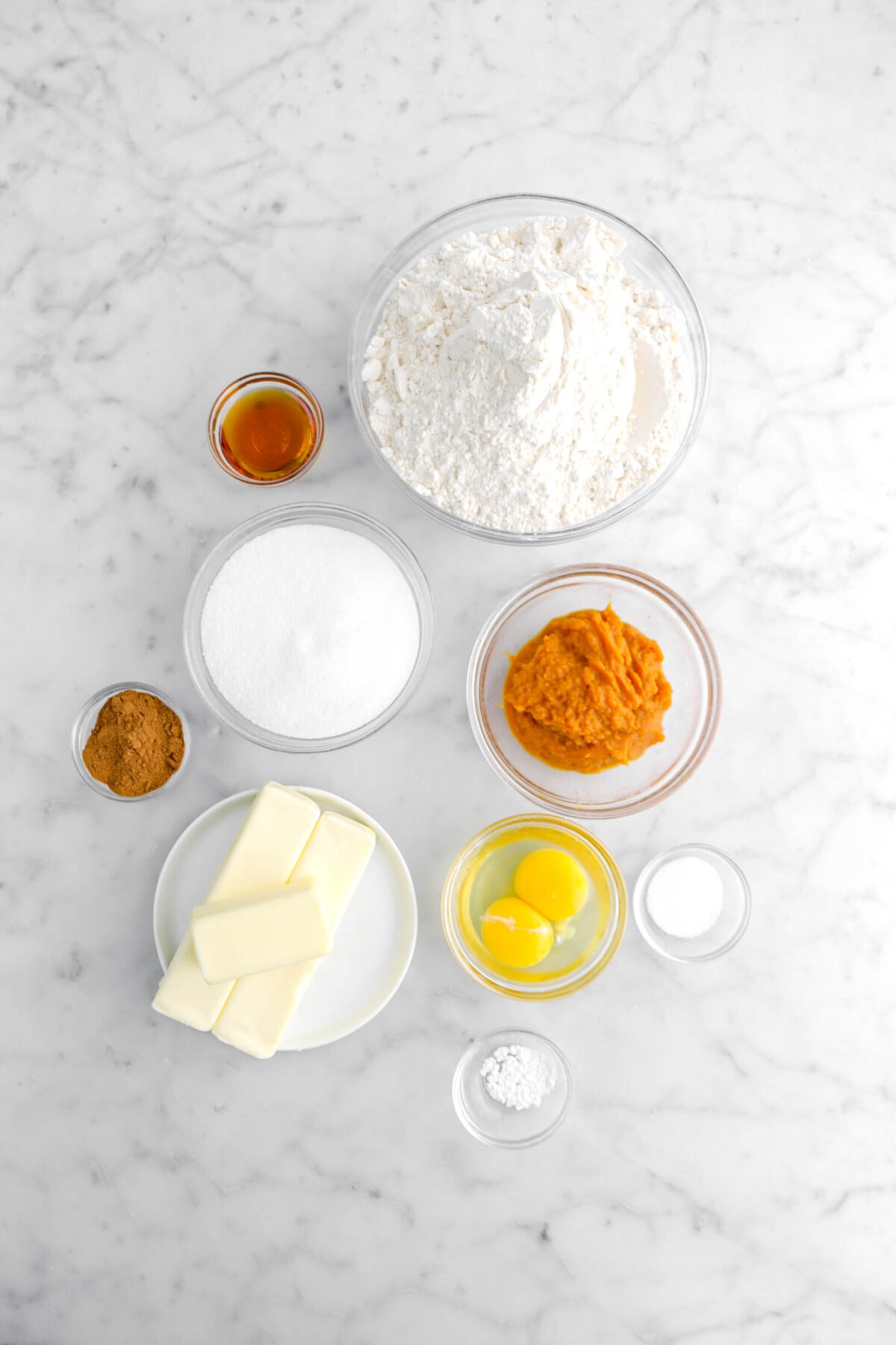 vanilla, flour, sugar, baking powder, pumpkin pie spice, pumpkin puree, eggs, butter, and salt on marble surface.