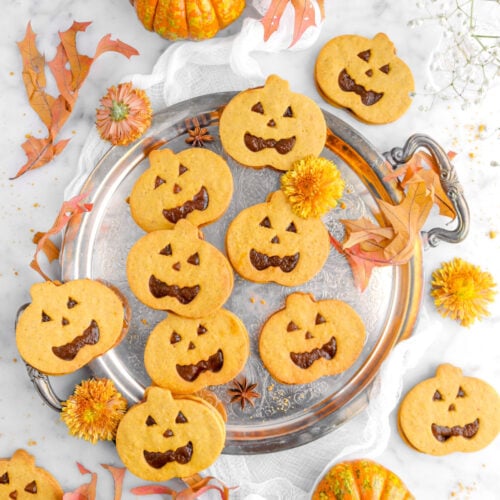 overhead shot of pumpkin jack o'lantern sandwich cookies on silver tray with leaves, yellow chrysanthemums, and orange pumpkins around on marble surface.