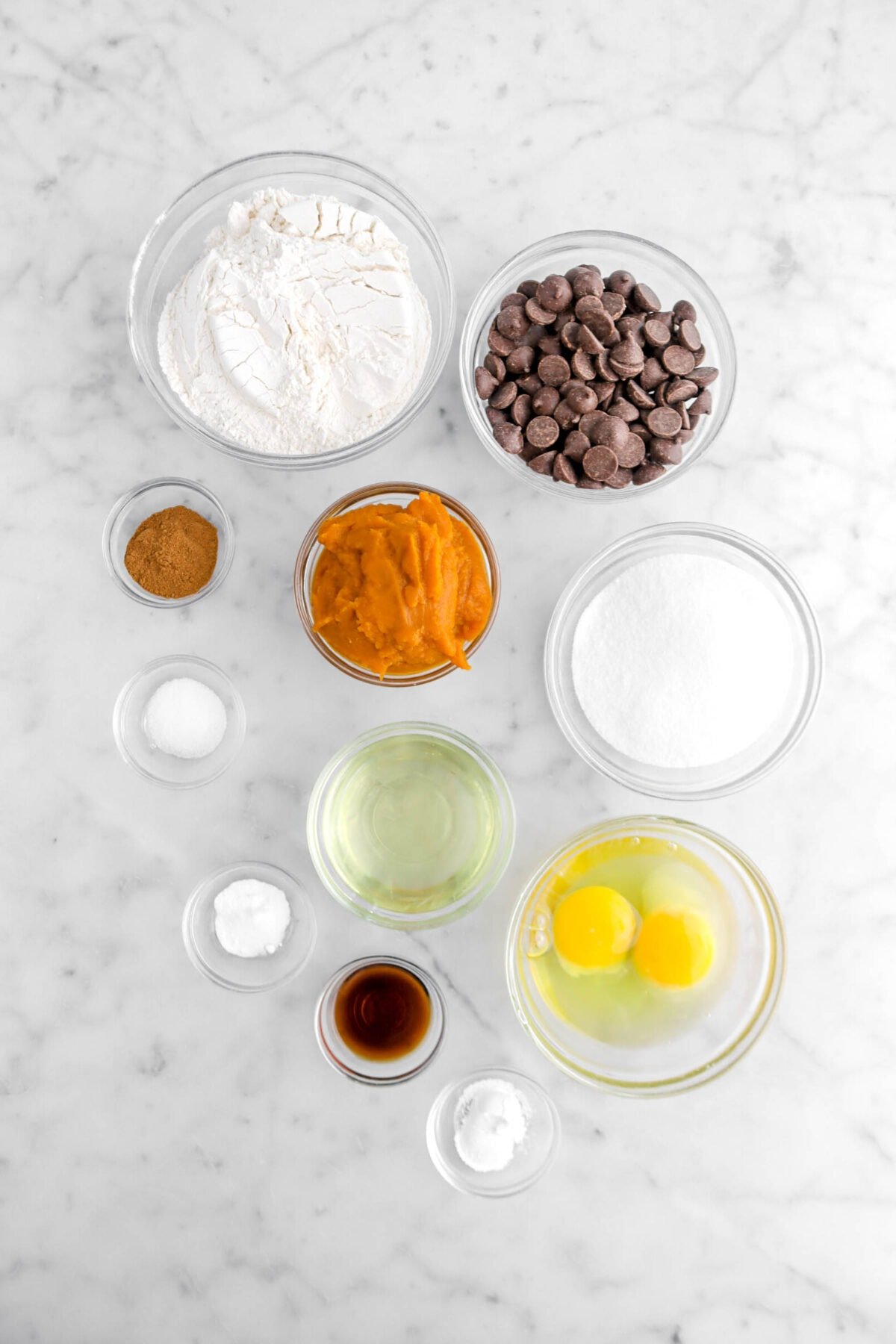 flour, chocolate chips, pumpkin pie spice, pumpkin purée, sugar, salt, baking soda, baking powder, vanilla, vegetable oil, and eggs on marble surface.