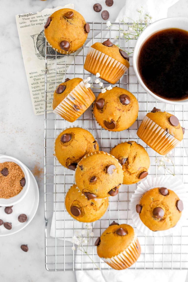 eleven pumpkin chocolate chip muffins on wire cooling rack with one stacked on top of three other muffins, a white napkin, a cup of coffee, and white flowers around on marble surface with chocolate chips and bowl of bowl sugar beside.