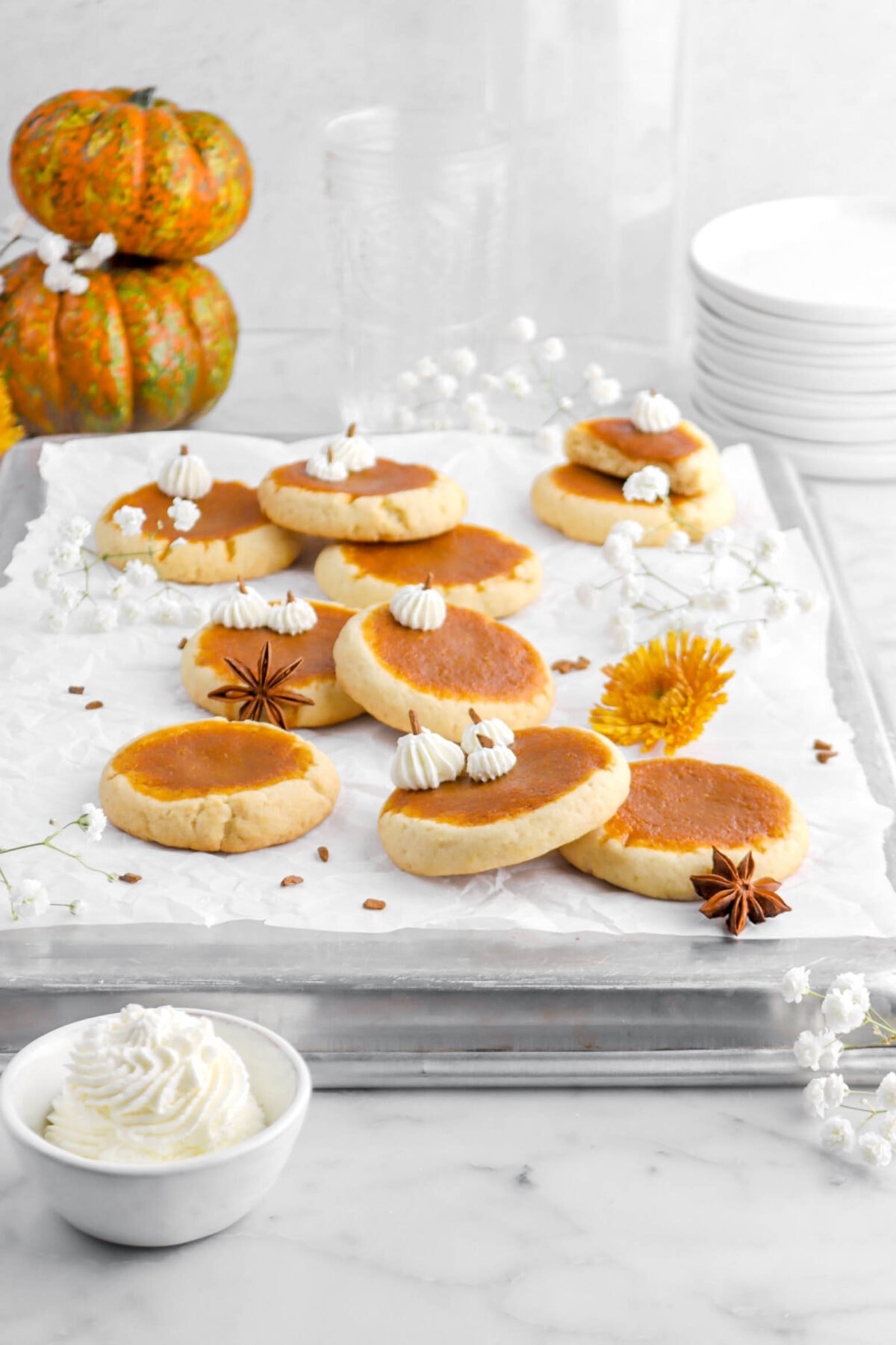 pumpkin pie cookies on upside down sheet pan with bowl of whipped cream, white flowers, stack of pumpkins, and plates behind.