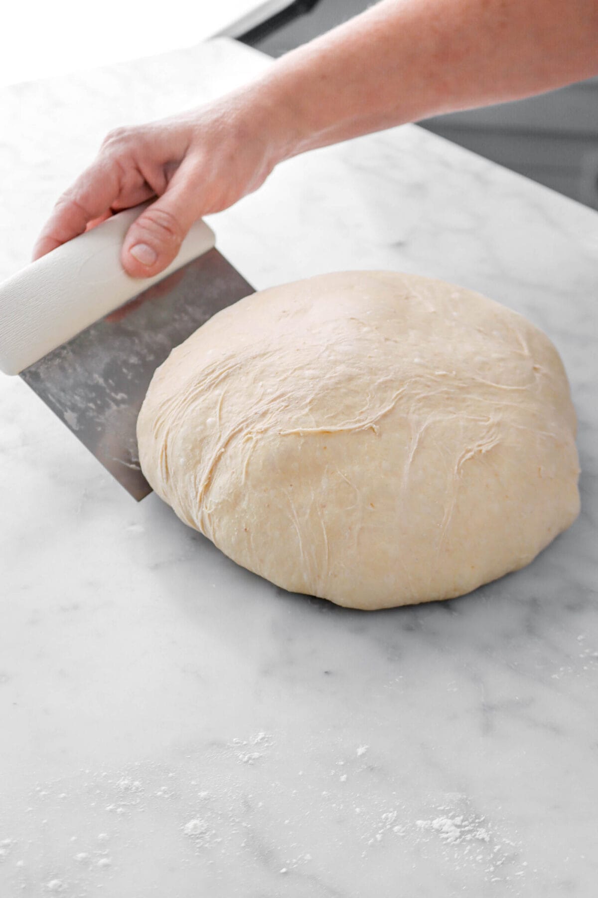 dough being shaped into a boule with bench scraper on marble surface.