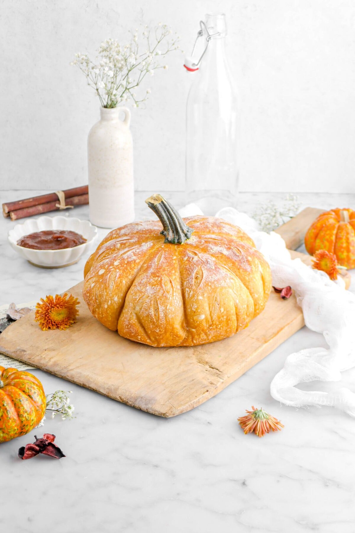 pulled back angled shot of pumpkin bread on wood board with two more pumpkins around, orange mums, and a white cheesecloth around, a bowl of pumpkin butter, stack of sticks, and a vase behind.