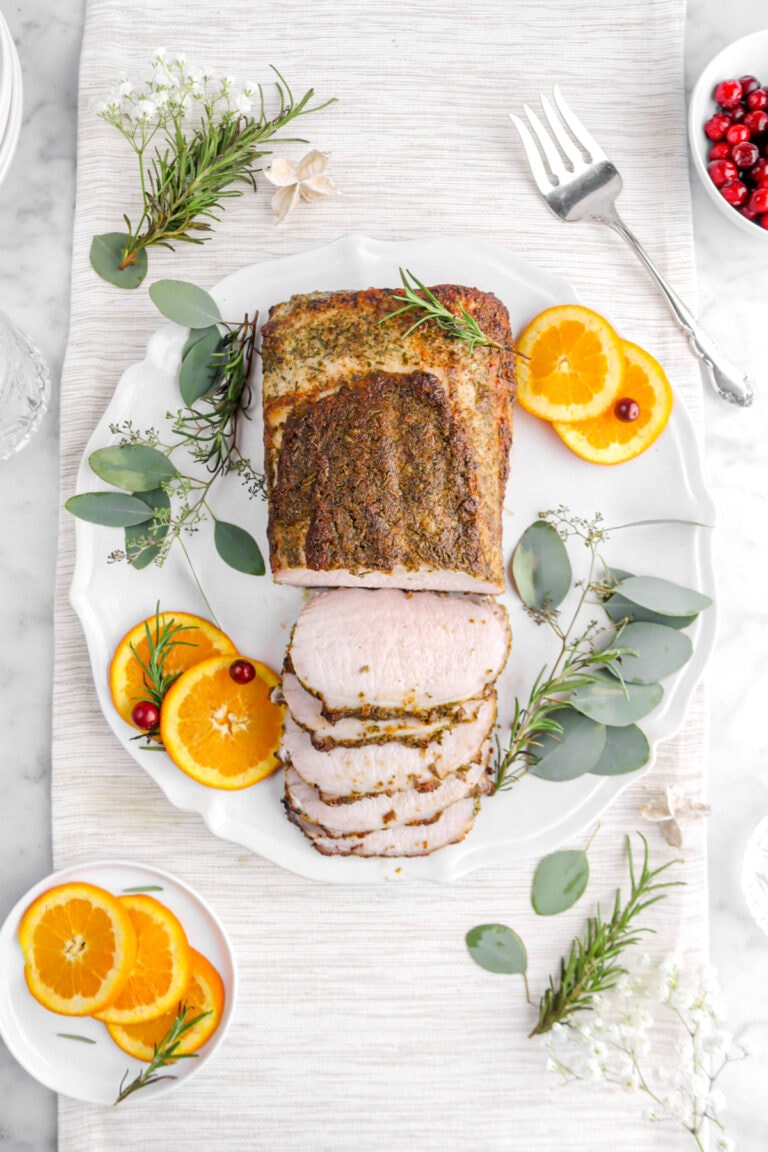 overhead shot of pork tenderloin with five slices laying in front on round platter with orange slices, rosemary sprigs, eucalyptus leaves, and cranberries around.