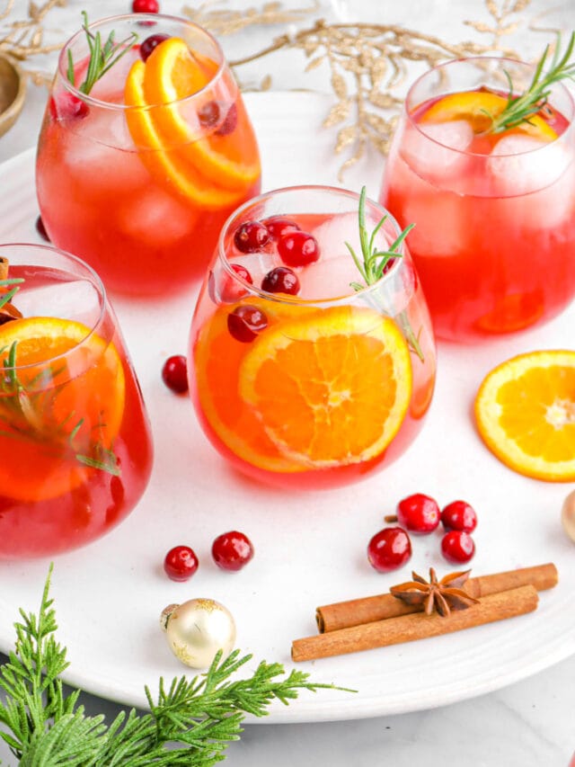 angled photo of four glasses of punch on white platter with whole spices, cranberries, a small ornament, christmas greenery, and gold leaf garland behind.