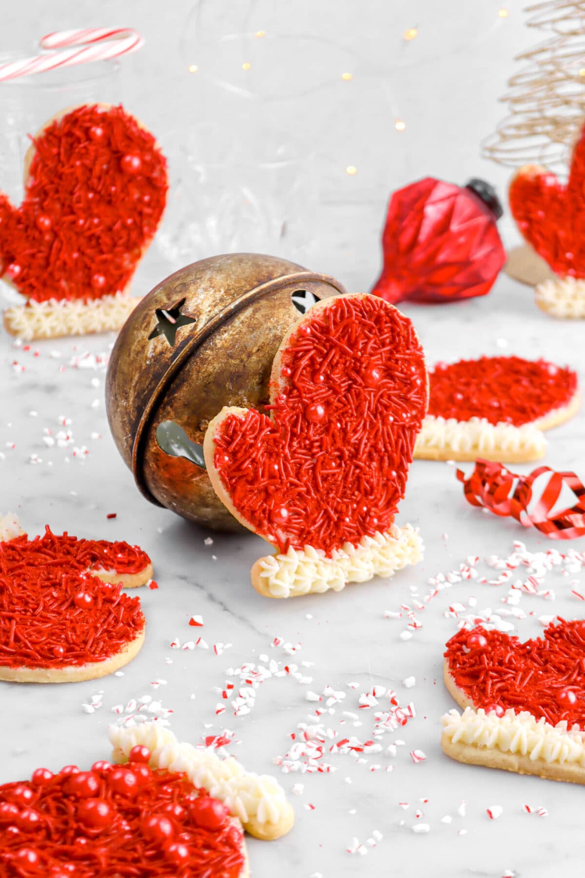 seven glove shaped cookies, with one leaning against a red bell in the middle on marble surface with crushed peppermint around.