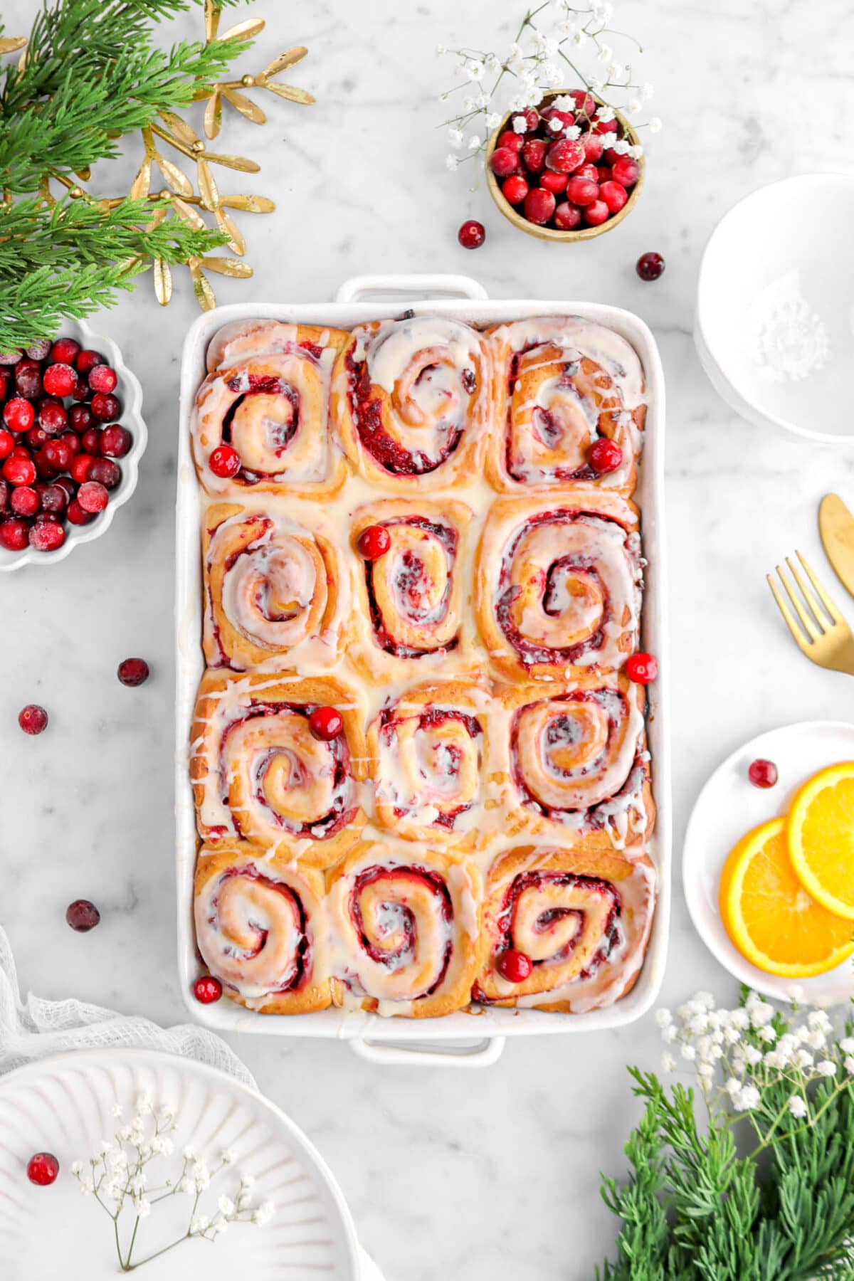 cranberry sweet rolls in rectangular casserole with cranberries around, a plate of orange slices, and christmas greenery around.