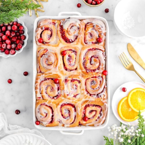 overhead shot of sweet rolls with cranberries around, a plate beside, christmas greenery, two slices of orange, and gold utensils beside.