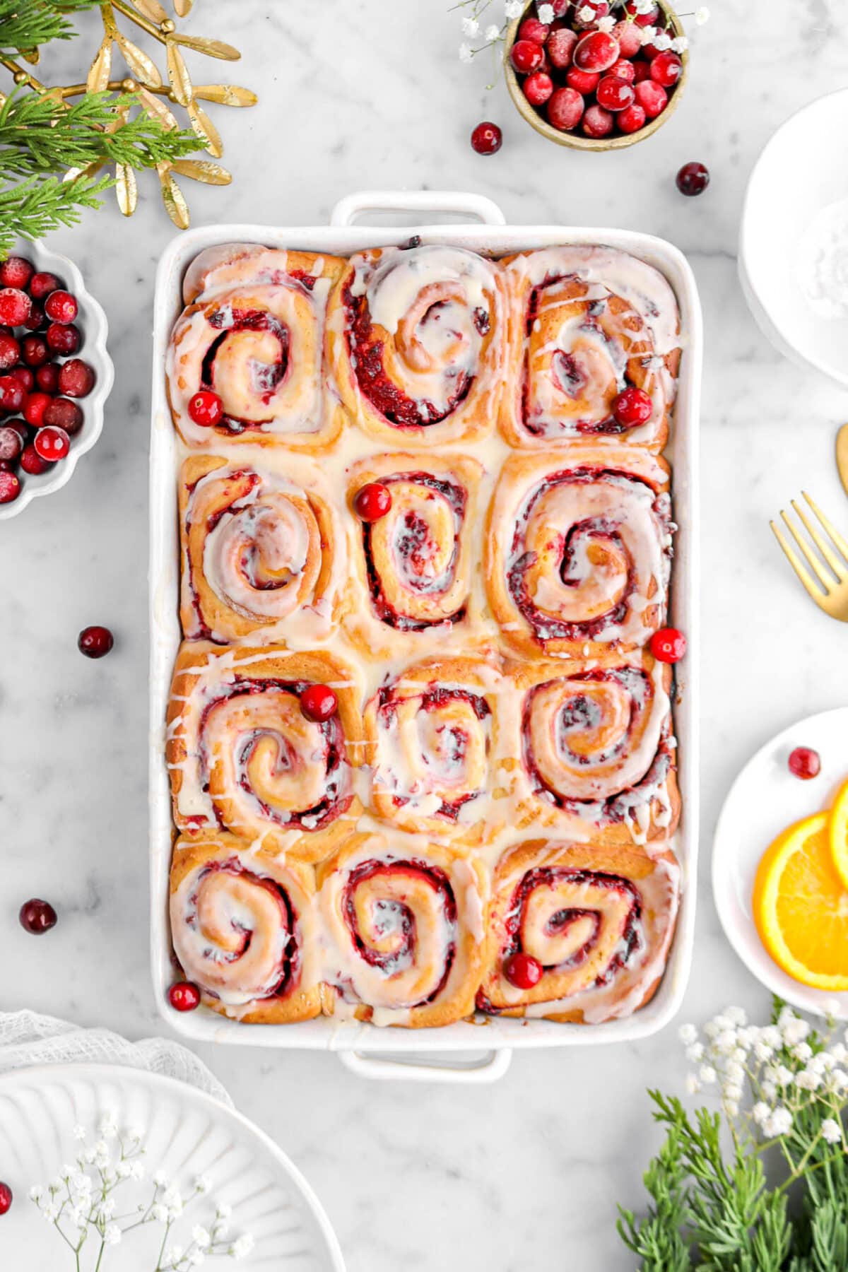 twelve cranberry sweet rolls in casserole with frozen cranberries around, white flowers, christmas greenery, and a plate