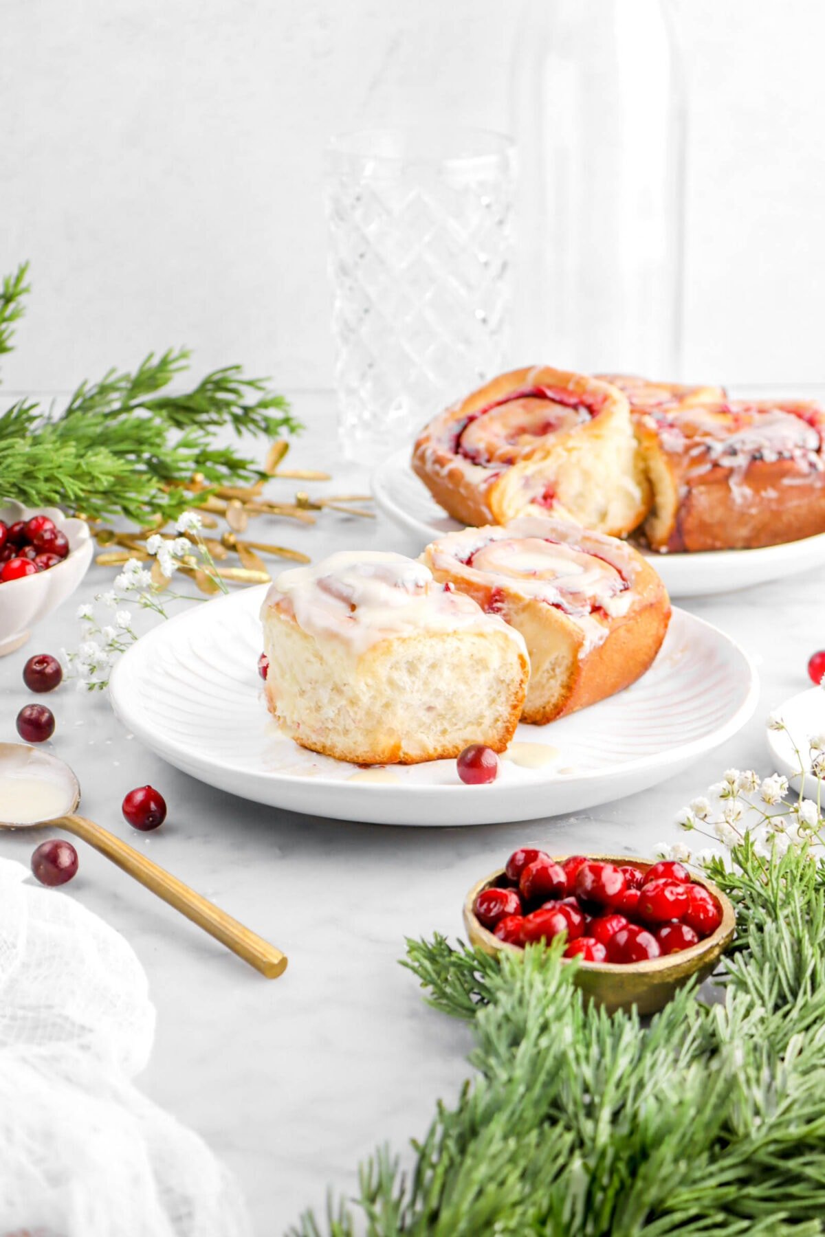 plate of two sweet rolls on white plate with greenery around and cranberries around on marble surface.