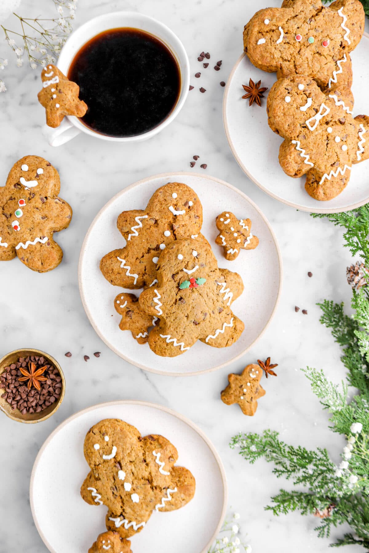 three white plates, each containing gingerbread men shaped scones, with three more scones around on marble surface, with mug of coffee, gold bowl of chocolate chips with star anise pod, and green garland beside.