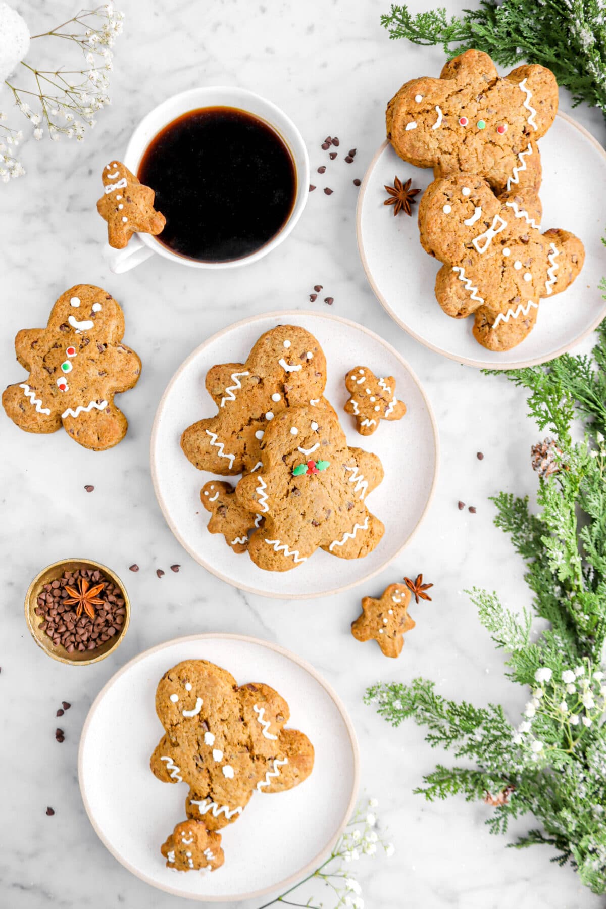 pulled back overhead shot of three plates with gingerbread scones on marble surface with greenery beside, gold bowl of mini chocolate chips, and white flowers around.