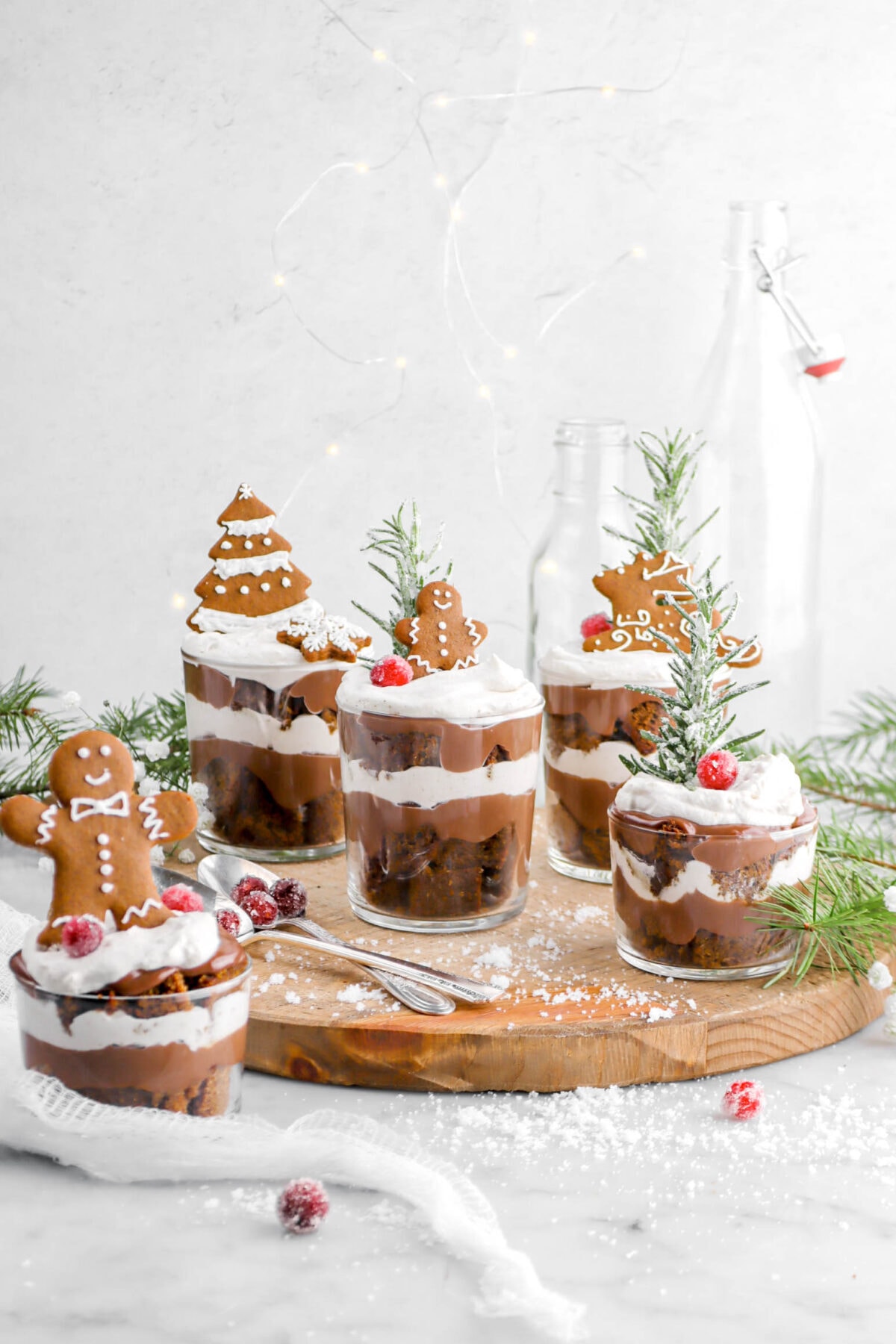 pulled back shot of mini gingerbread trifles on wood board with sugared cranberries, two spoons, pine branches, and a white cheese cloth around.