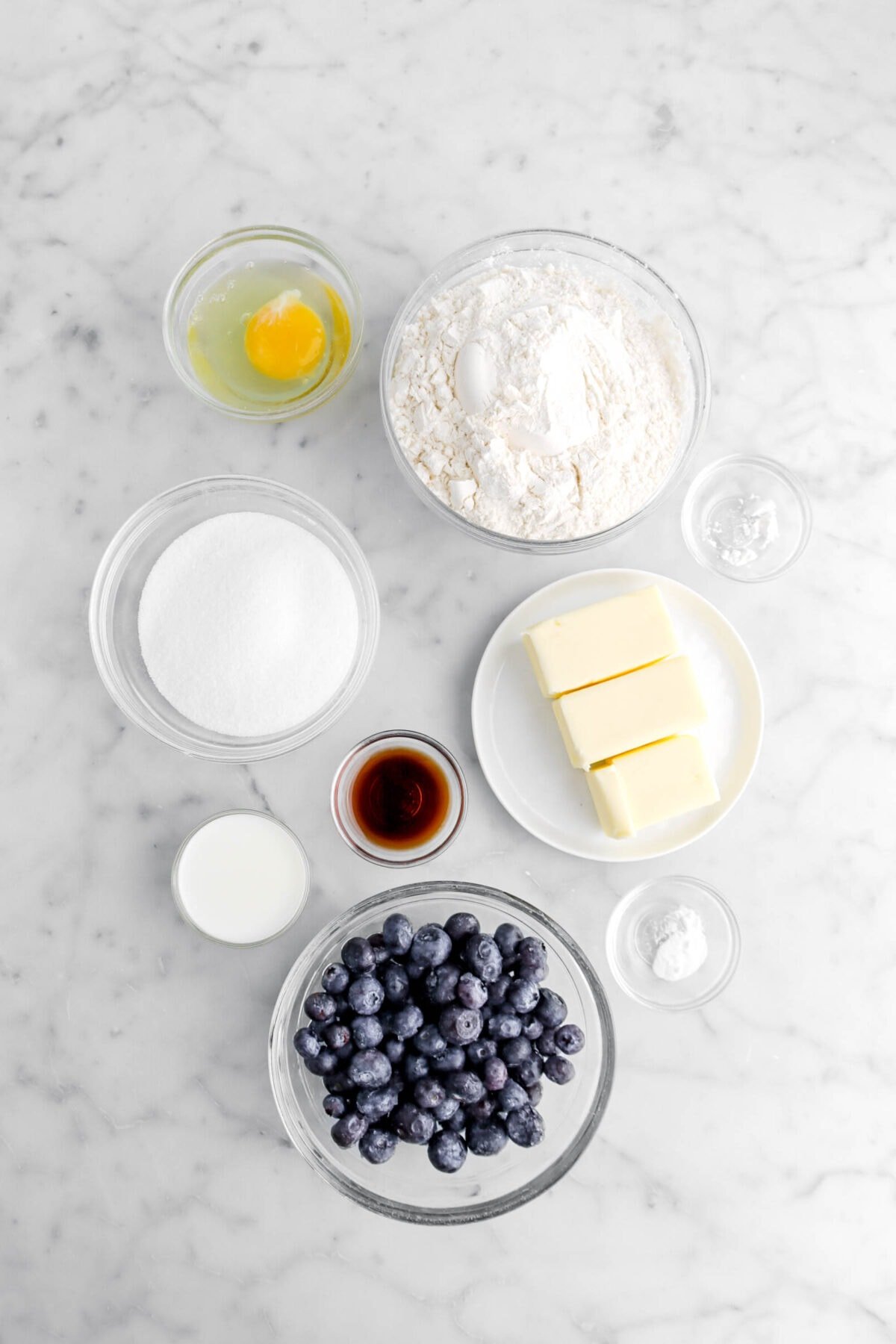 egg, flour, sugar, baking powder, butter, vanilla, milk, baking soda, and fresh blueberries on marble surface.