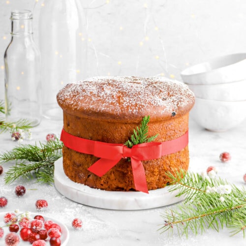 panettone dusted lightly with powdered sugar and wrapped in red ribbon with small bow on marble stand with pine greenery around and sugared cranberries on marble surface with stack of coffee mugs and string lights behind.