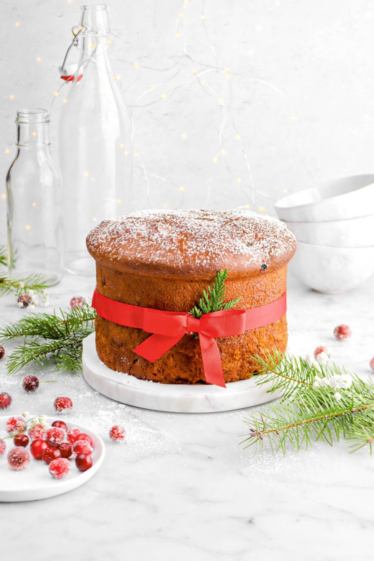 panettone dusted lightly with powdered sugar and wrapped in red ribbon with small bow on marble stand with pine greenery around and sugared cranberries on marble surface with stack of coffee mugs and string lights behind.