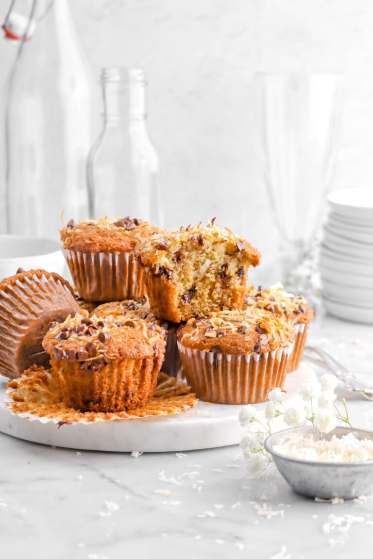 muffins stacked on marble tray with one muffin missing a bite, with bowl of shredded coconut and white flower beside.
