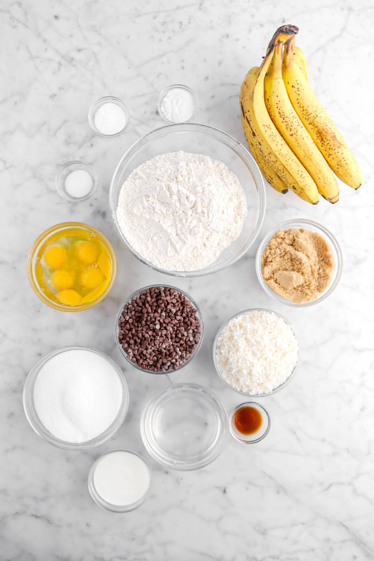 baking powder, baking soda, salt, flour, bananas, eggs, chocolate chips, brown sugar, coconut, vanilla, coconut oil, sugar, and buttermilk on marble surface.